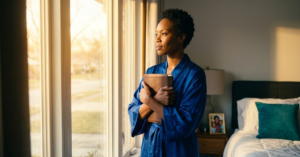 Your Worth Isn't Tied to Your Spiritual Streak Black woman in cobalt blue satin robe holding Bible pressed to her chest at bedroom window, expression of relief and peace, gold morning light